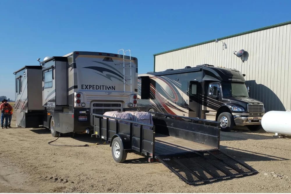 Two large RVs and a trailer are parked on a dirt lot next to a building, with two people in the background.