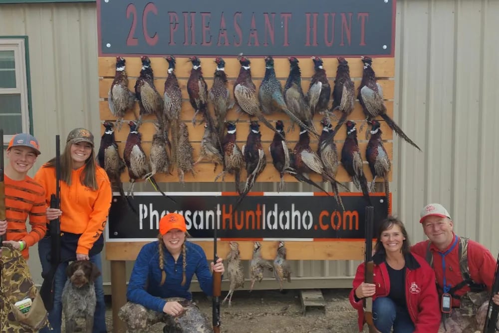A group of hunters and a dog pose with harvested ring-necked pheasants hanging on a wooden board.