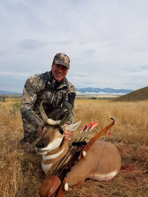 A hunter in camouflage kneels next to a harvested pronghorn with a bow and arrows in a grassy field.