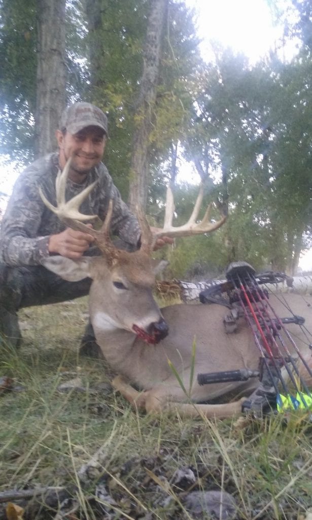 A hunter in camouflage kneels next to a harvested whitetail deer, holding its antlers, with a bow resting on the deer's body.