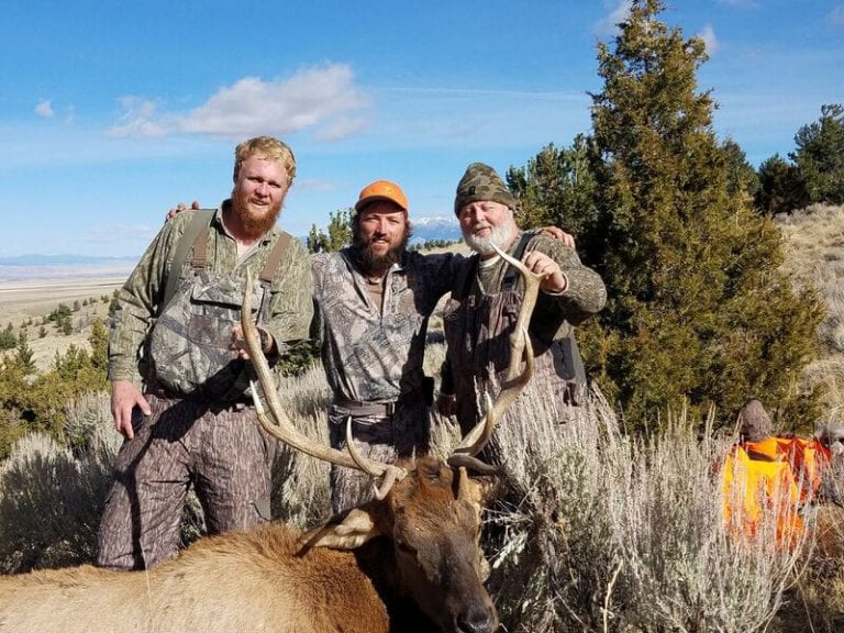 Three hunters pose with a harvested elk in a field.
