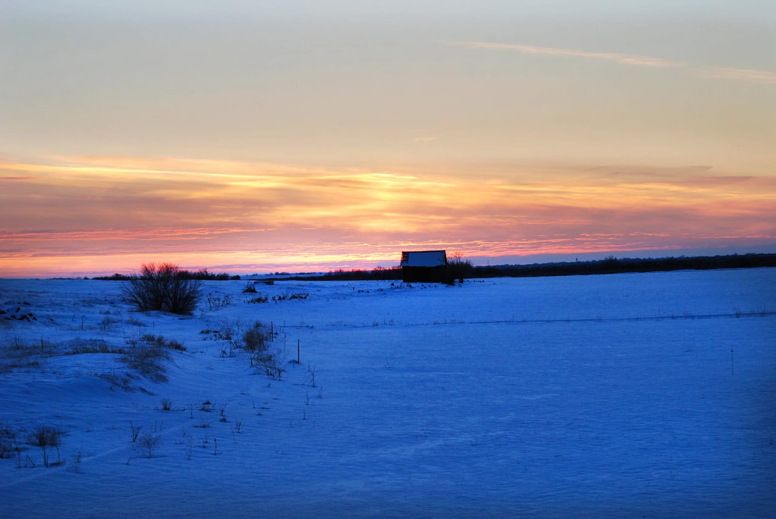 A snowy field at sunset with a building in the distance.