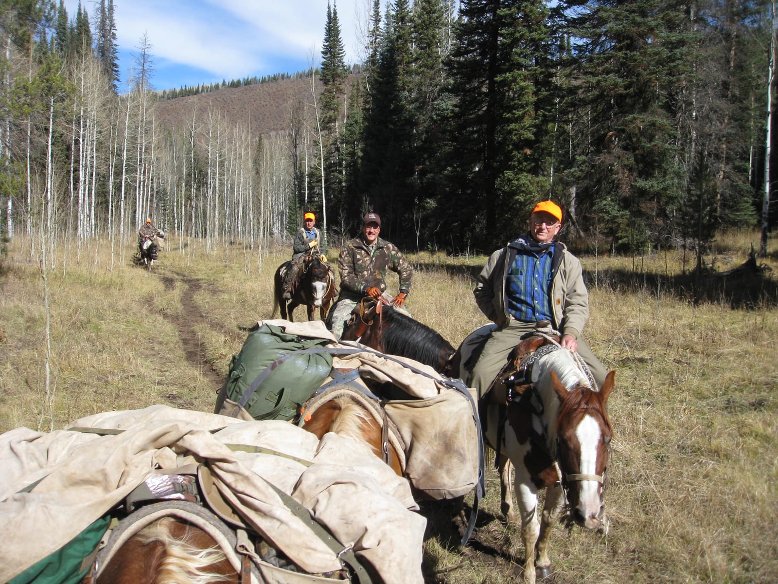 Several men on horseback with packed gear ride along a dirt path through a forest.