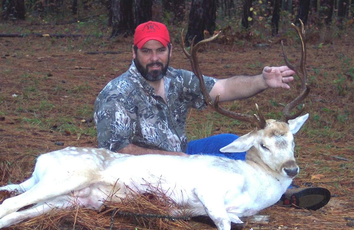 A hunter in a red hat and camouflage shirt poses with a harvested white fallow deer.