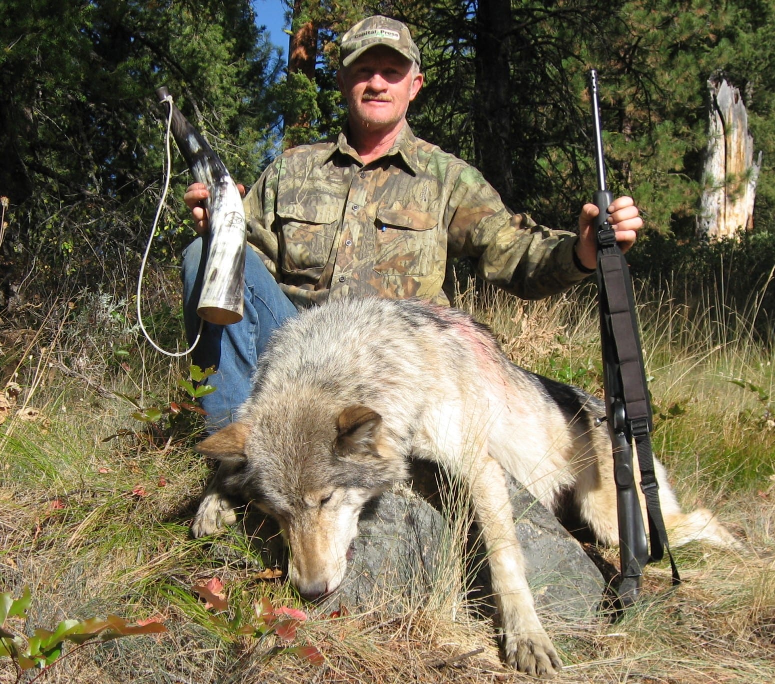 A hunter poses with a harvested wolf and a rifle in a wooded area.