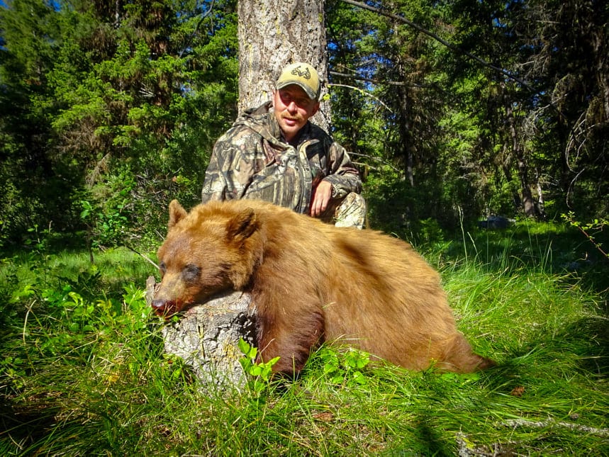 A hunter in camouflage poses with a harvested black bear next to a tree in a forest.