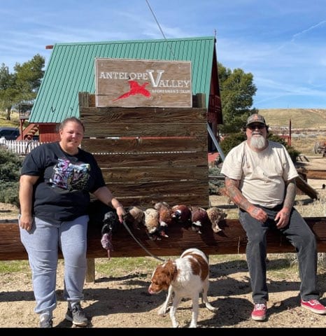 Two people and a dog pose with harvested chukar partridges in front of an Antelope Valley sign.