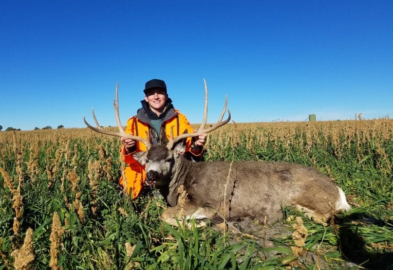A hunter poses with a harvested mule deer in a field under a clear blue sky.