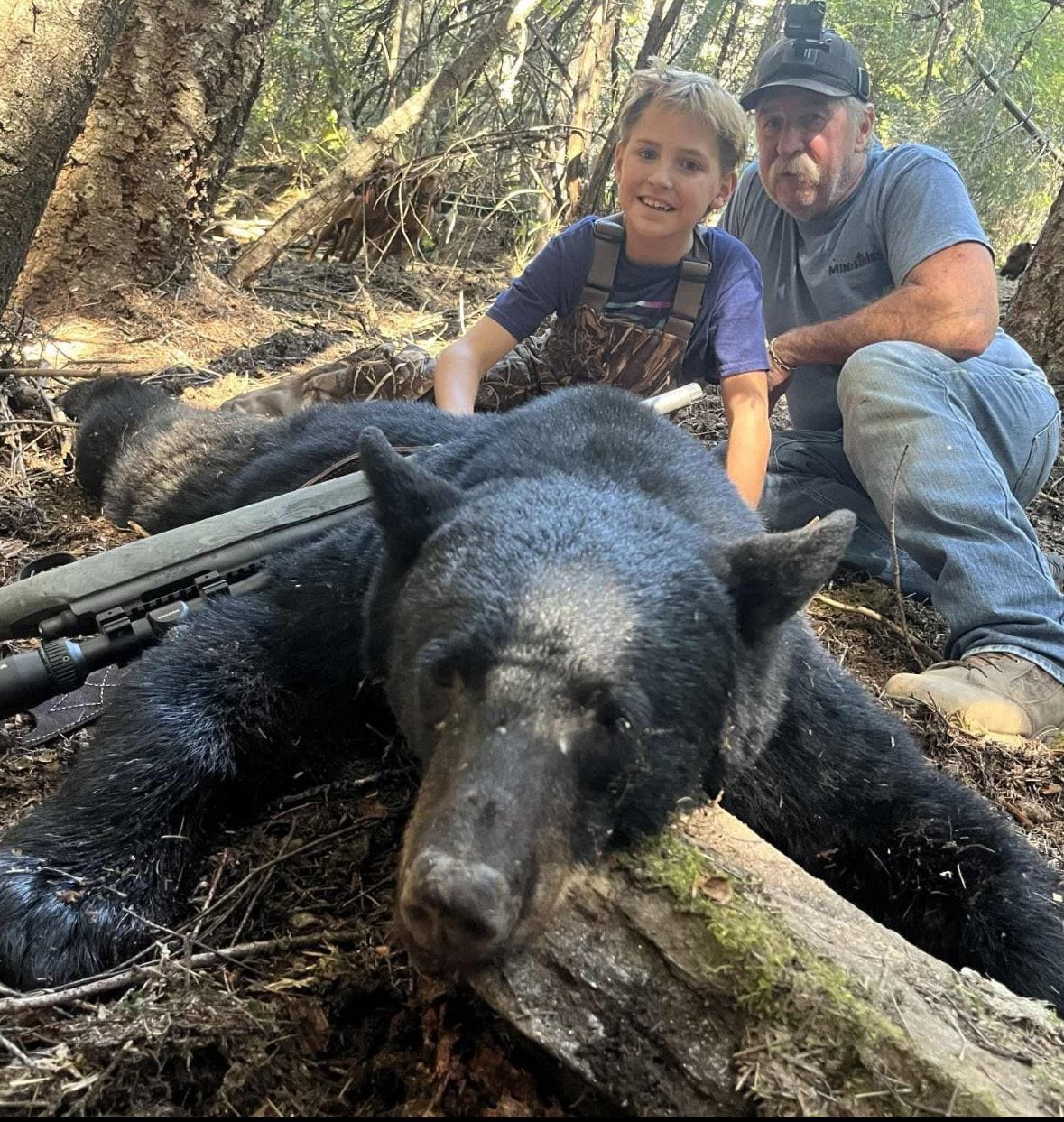 A hunter and a child pose with a harvested black bear in a forest.