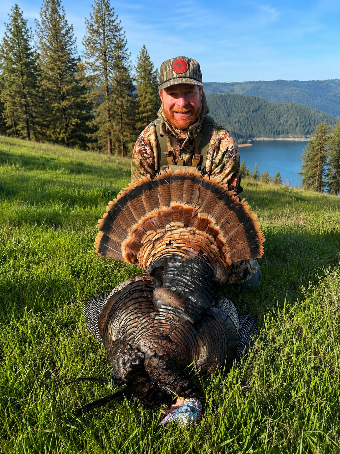 A hunter poses with a harvested wild turkey in a grassy field with a lake and mountains in the background.