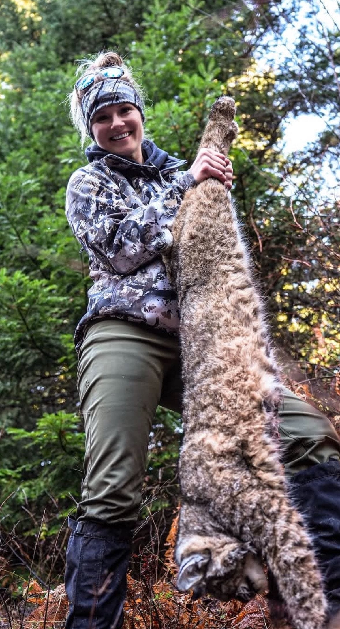A woman in camouflage holds a harvested bobcat in a forest.