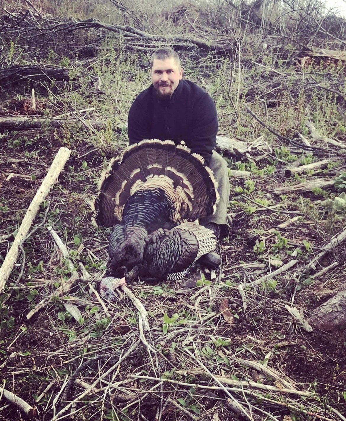 A man kneels with two harvested wild turkeys in a brushy outdoor setting.