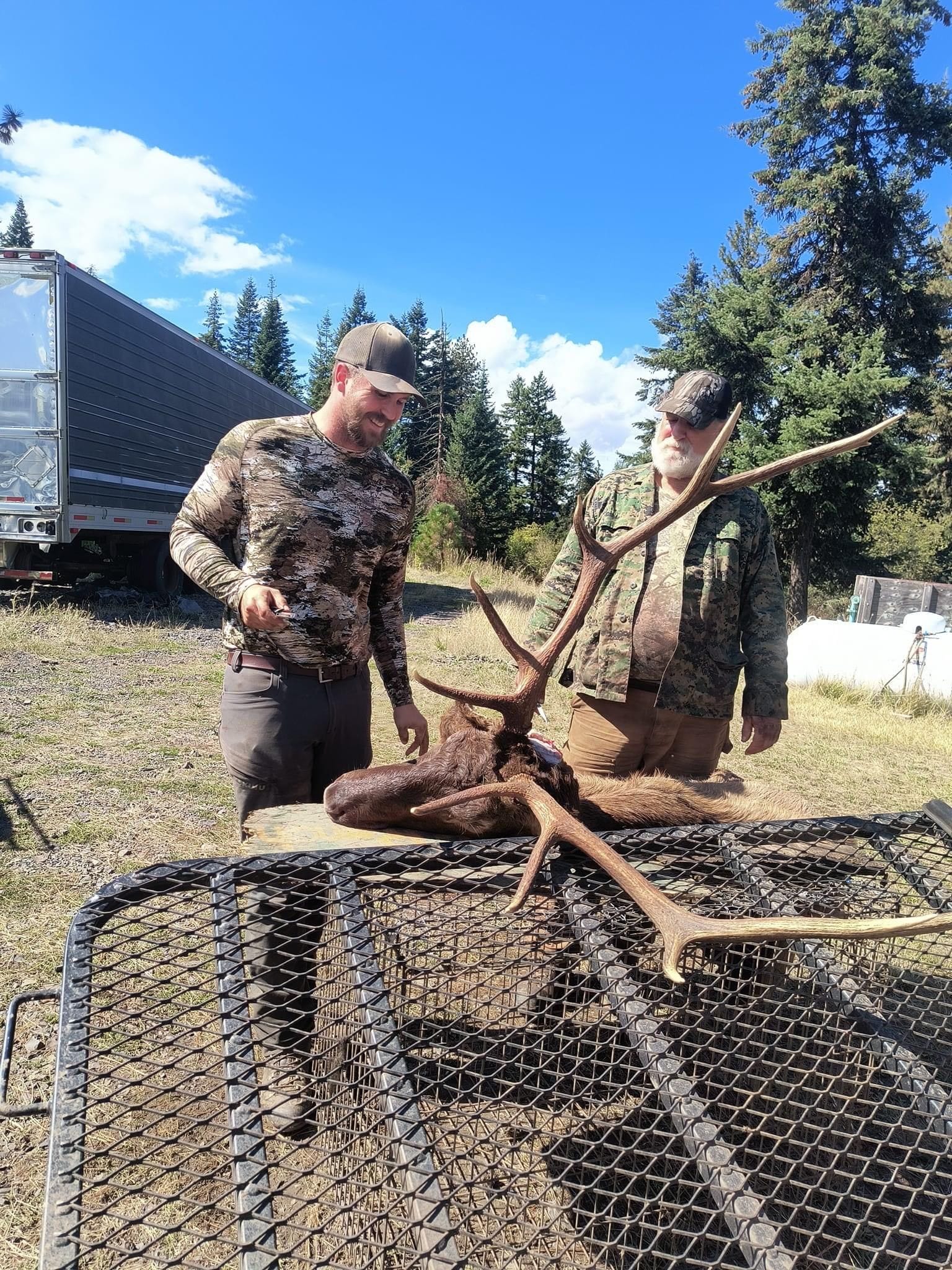 Two men stand next to a harvested elk head with antlers.