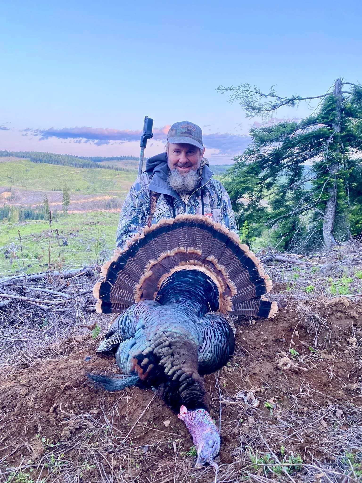 A man in camouflage poses with a harvested wild turkey in a field.