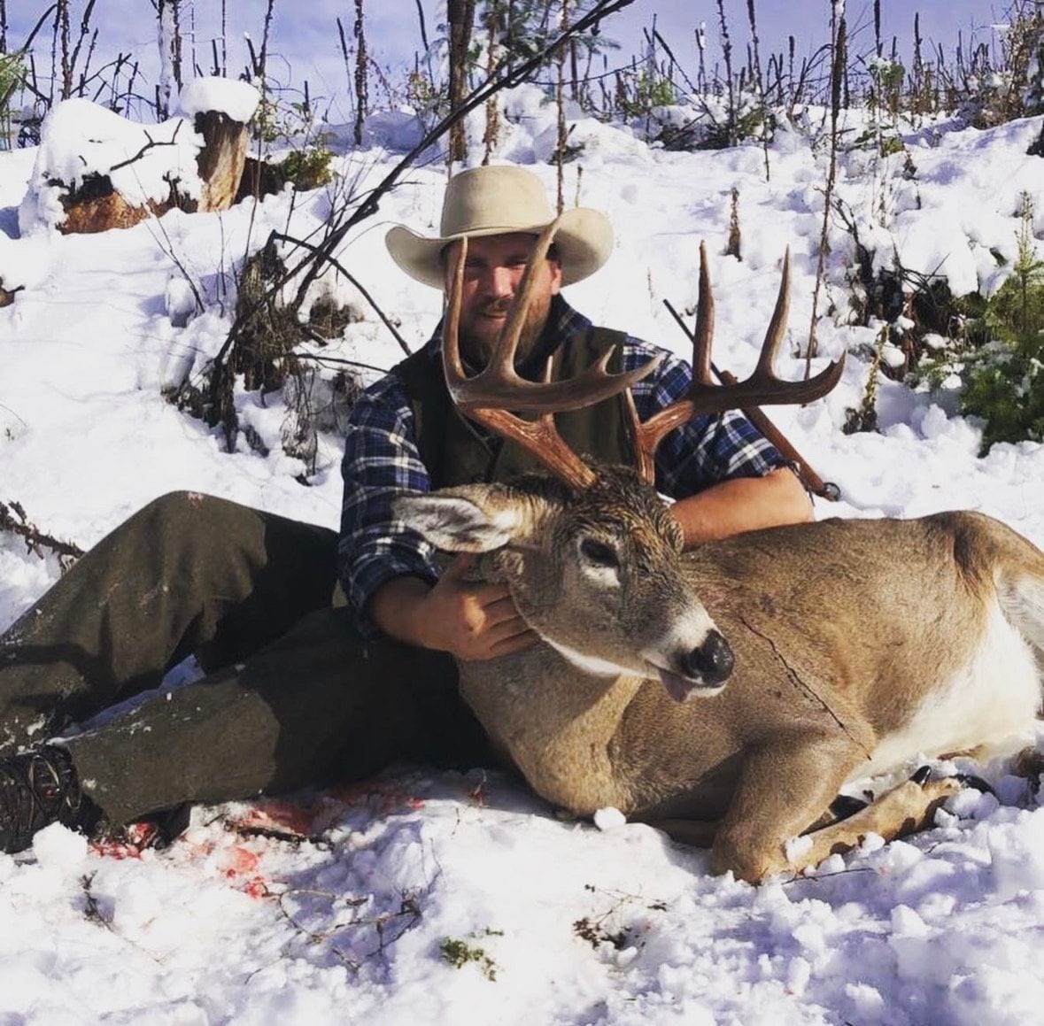 A hunter in a cowboy hat poses with a harvested whitetail deer in the snow.