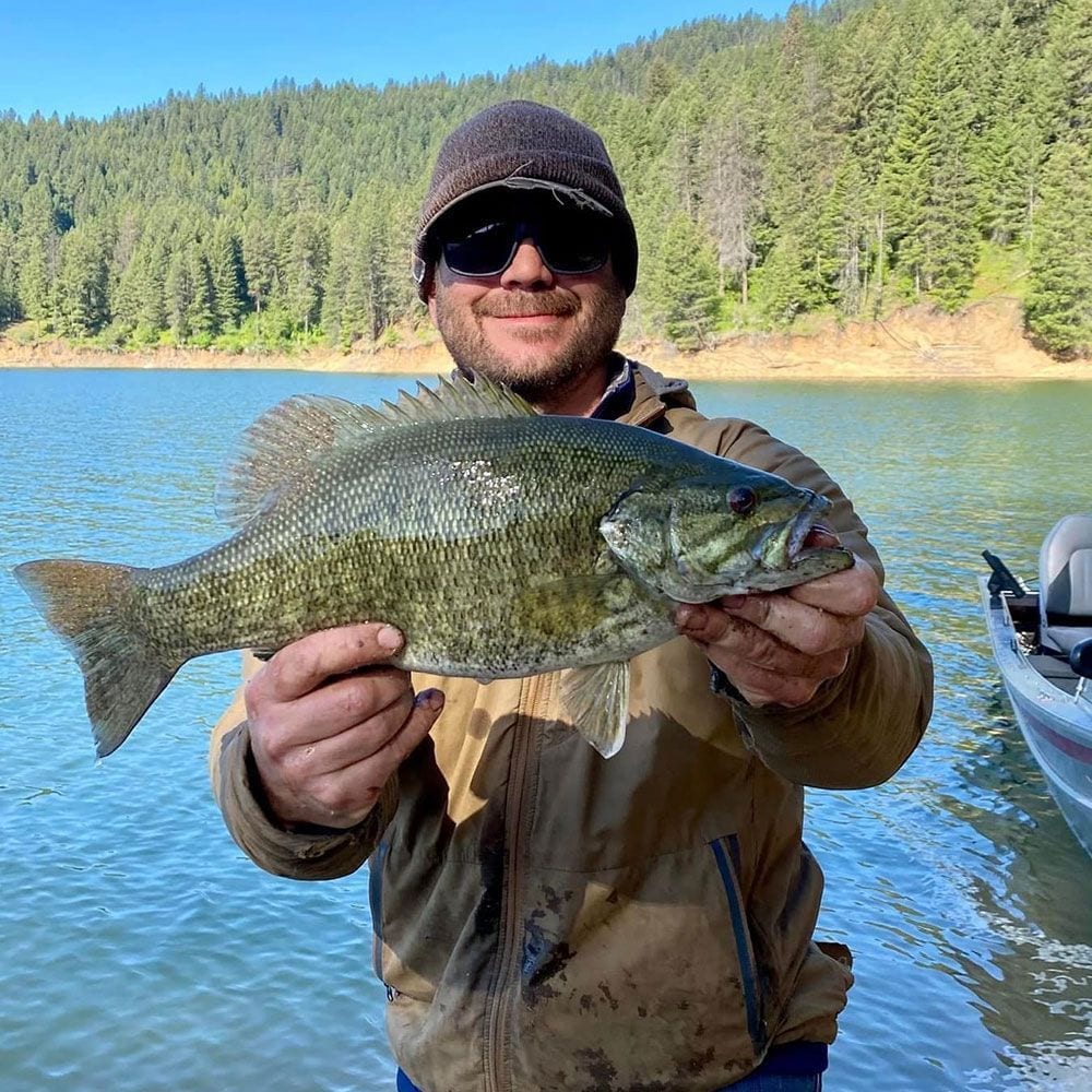 A man holds a fish in front of a lake and forested hills.
