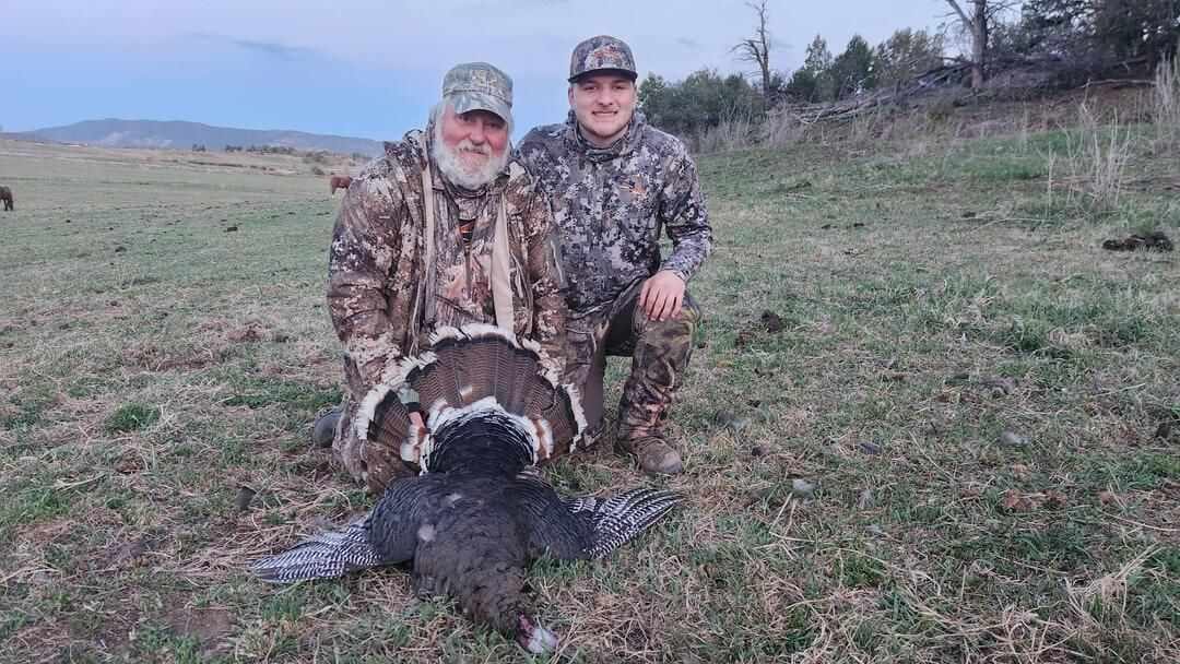Two hunters pose with a harvested wild turkey in a grassy field.