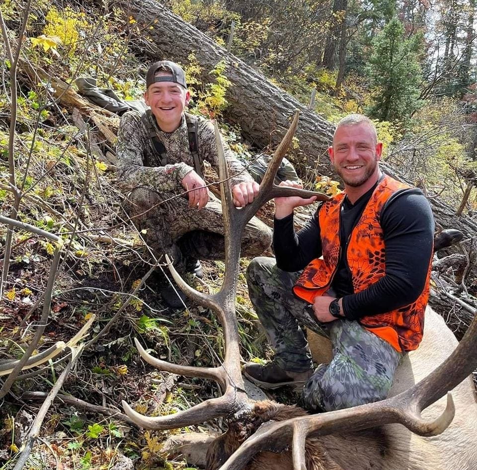 Two hunters pose with a harvested elk in a wooded area.