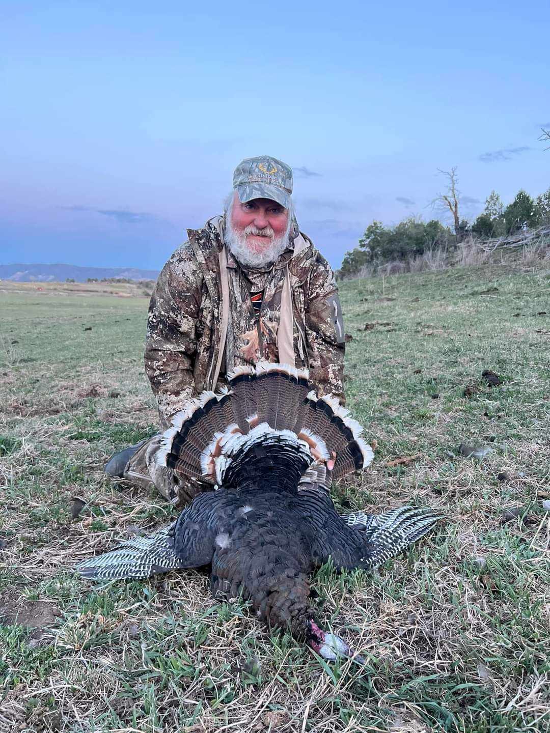 A hunter poses with a harvested wild turkey in a grassy field.