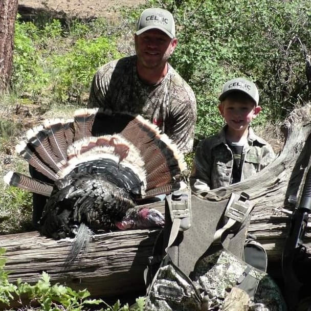 A man and a child pose with a harvested wild turkey on a log.