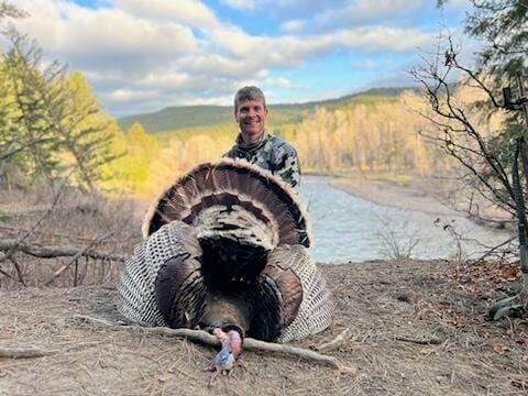 A man in camouflage poses with a harvested wild turkey, displaying its fanned tail feathers, with a river and forested hills in the background.
