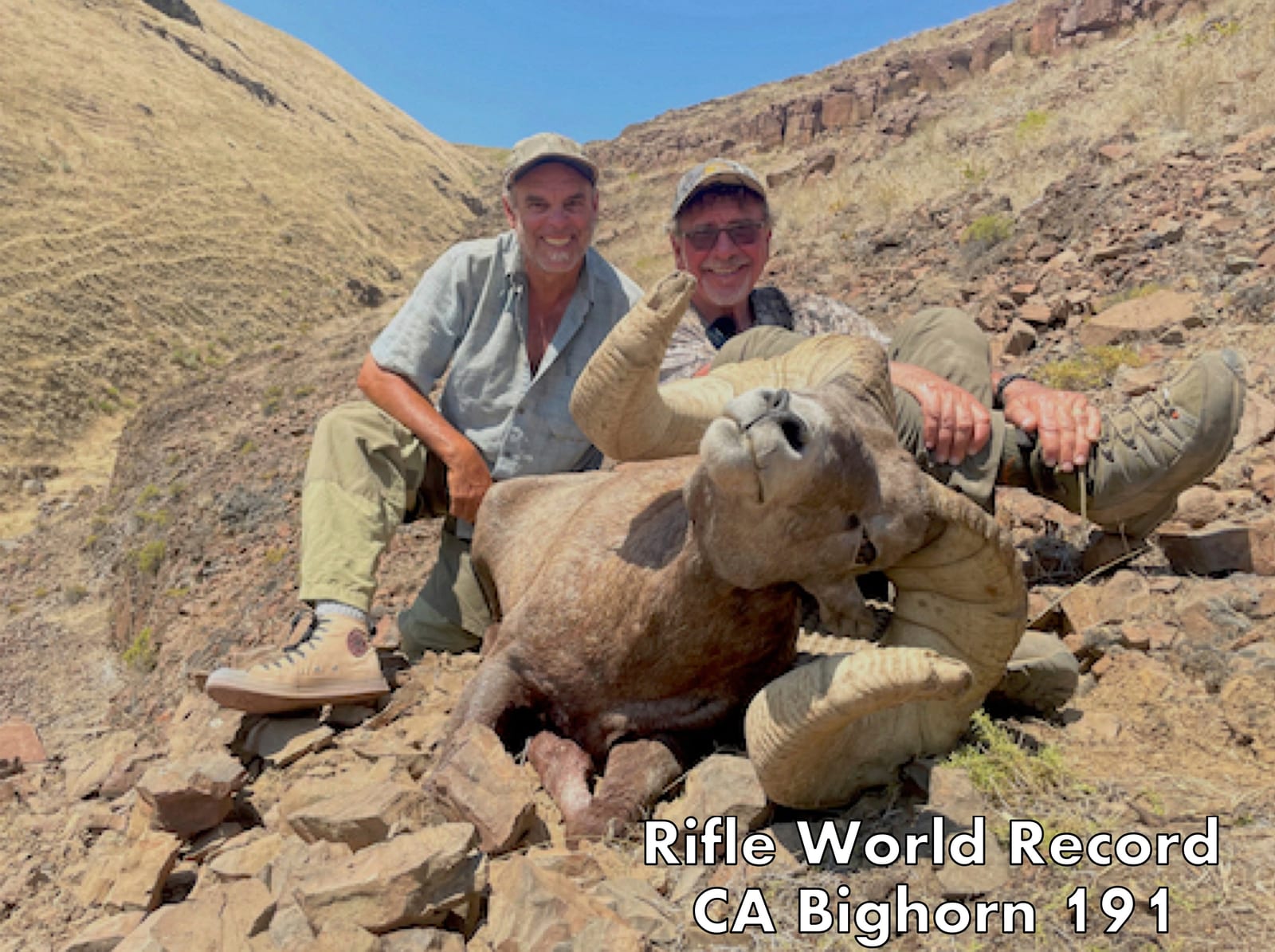 Two men pose with a harvested bighorn sheep, identified as a rifle world record California bighorn, in a rocky, arid landscape.