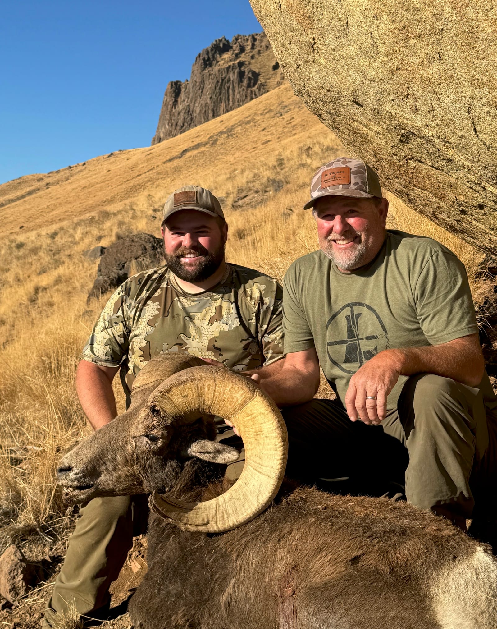 Two hunters pose with a harvested bighorn sheep in a mountainous, arid landscape.