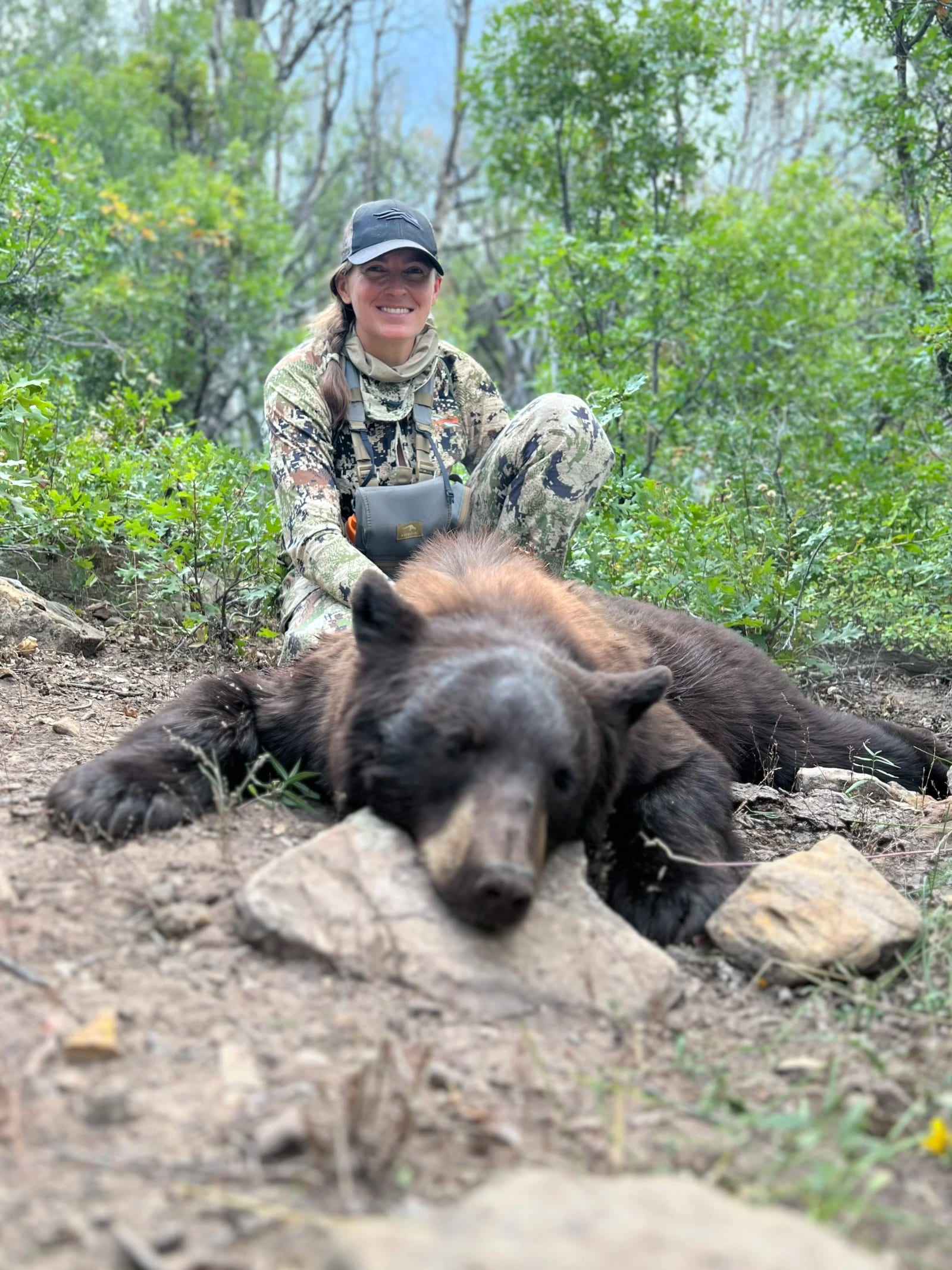 A woman in camouflage clothing and a cap kneels on the ground in a forest, smiling behind a harvested black bear with brown-tan fur, lying on the dirt among rocks and green foliage.