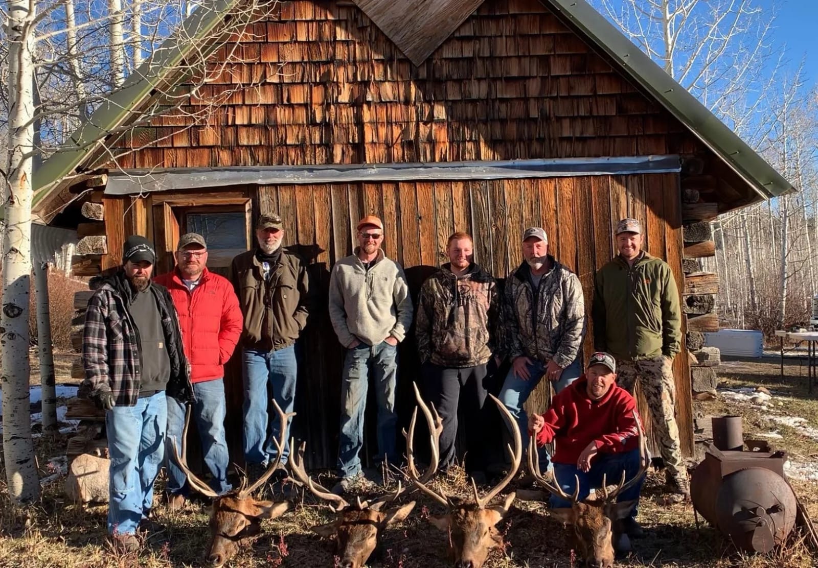 A group of nine men standing outdoors in front of a wooden cabin, with four large elk after a successful few days of hunting.