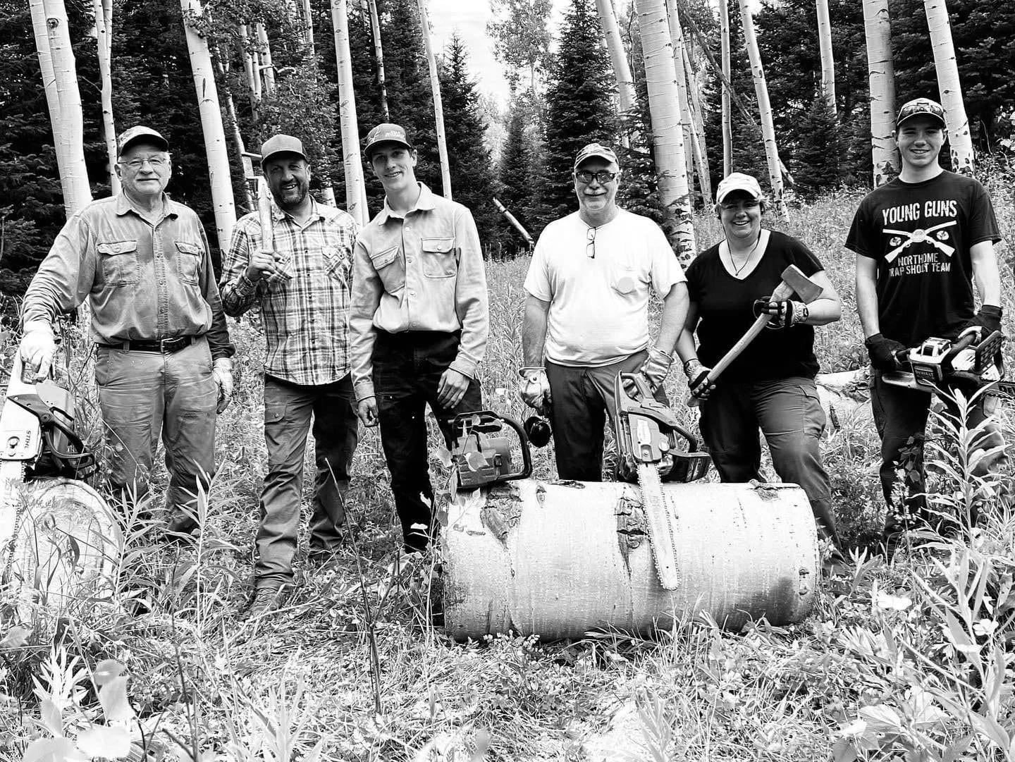 A group of six people, some holding chainsaws and axes, stand in a forest with aspen trees.