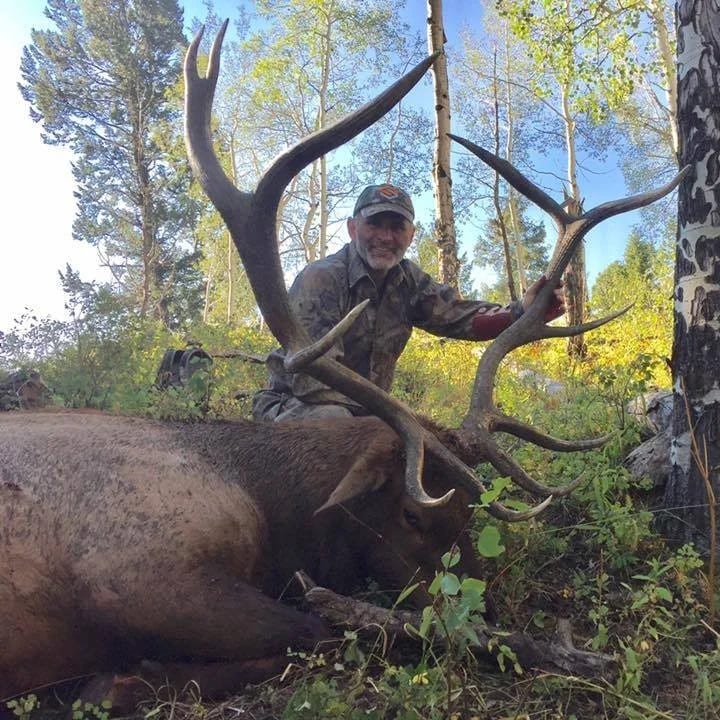 A hunter in camouflage poses with a harvested elk in a forest setting.