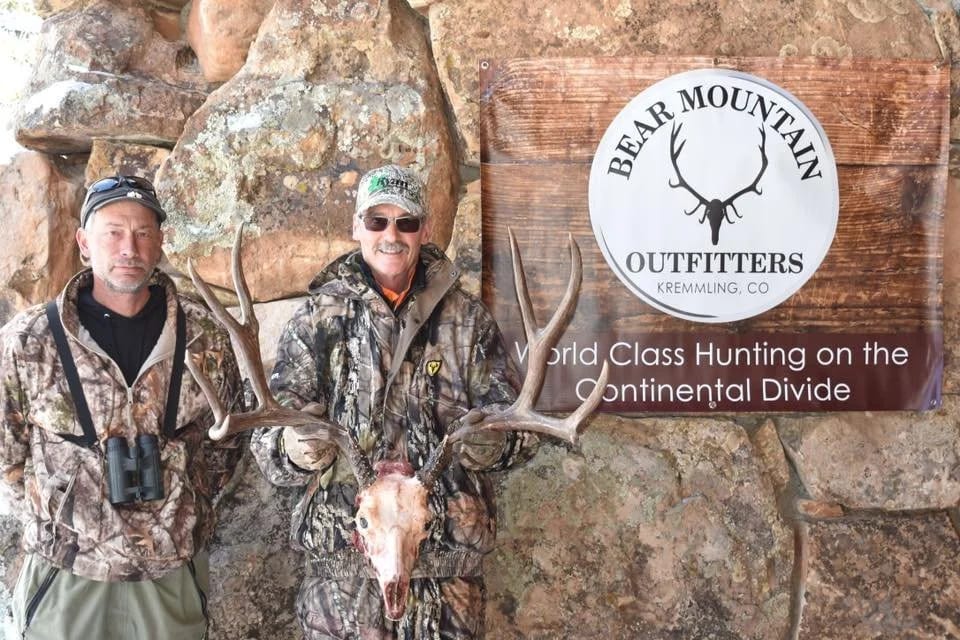 Two men in camouflage clothing pose with a harvested mule deer skull and antlers in front of a stone wall and a 'Bear Mountain Outfitters' sign.