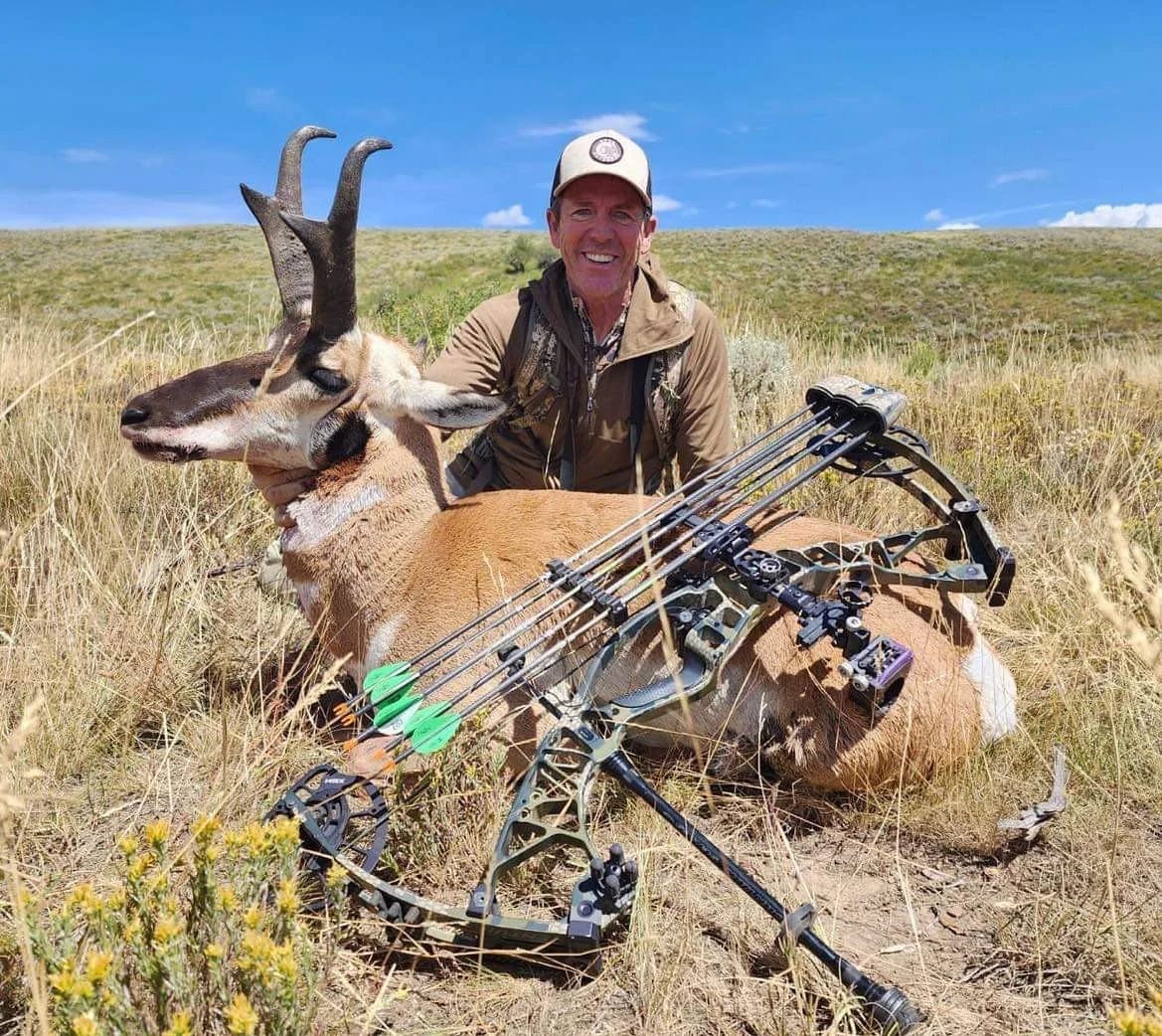 A hunter poses with a harvested pronghorn and a bow in a grassy field.