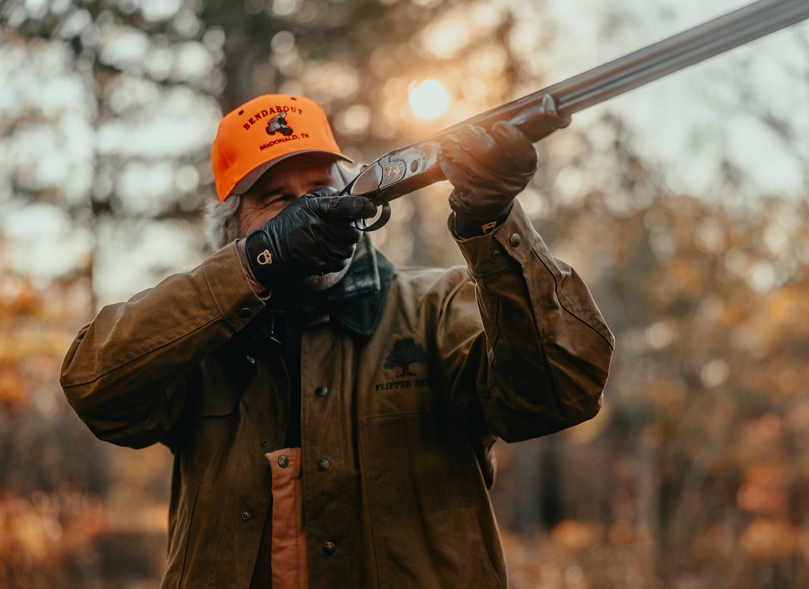 Man in a field wearing a blaze orange hat with shotgun raised.