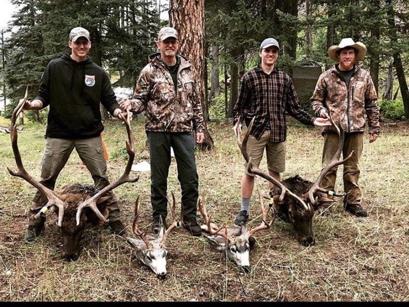 Four hunters pose with two harvested elk and one harvested mule deer in a forest.