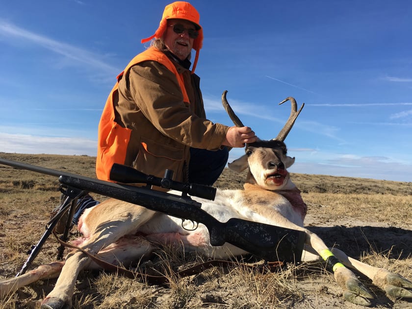 A hunter in an orange hat and vest poses with a harvested pronghorn in a dry, grassy field under a blue sky.