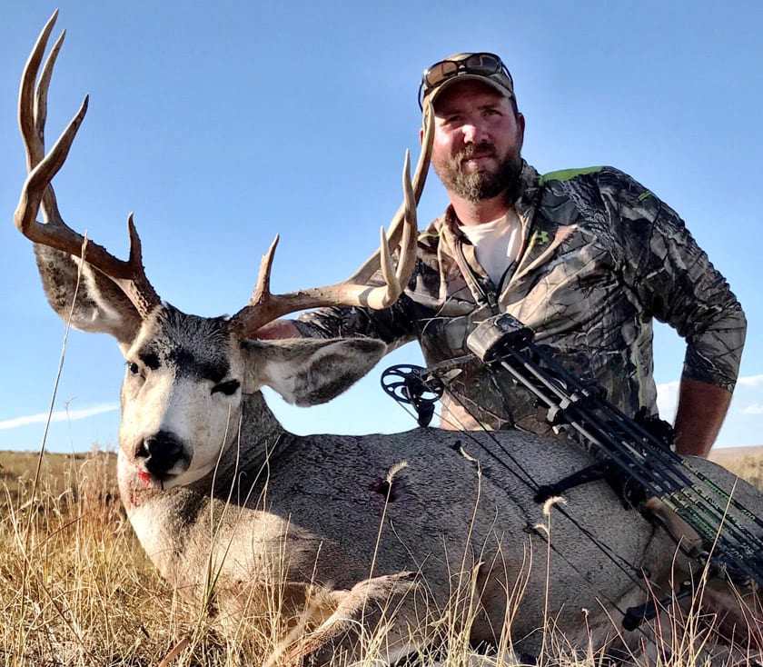 A hunter poses with a harvested mule deer and a bow in a grassy field.
