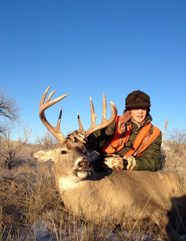 A hunter in an orange vest and cap poses with a harvested whitetail deer in a grassy field under a clear blue sky.