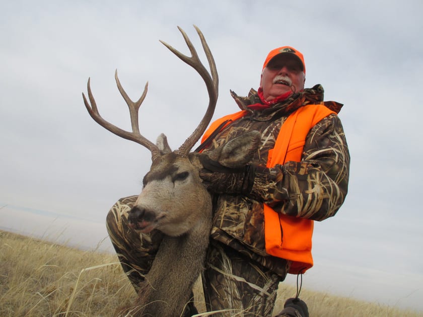 A hunter in camouflage and an orange vest poses with a harvested mule deer in a field.