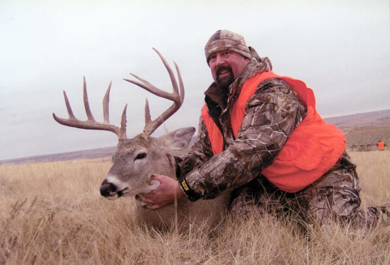 A hunter in camouflage and an orange vest poses with a harvested whitetail deer in a grassy field.
