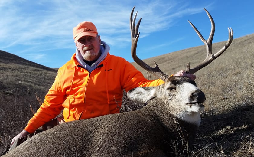 A hunter in an orange jacket and hat poses with a harvested mule deer on a grassy hillside.