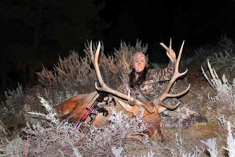 A hunter in camouflage poses with a harvested elk at night.