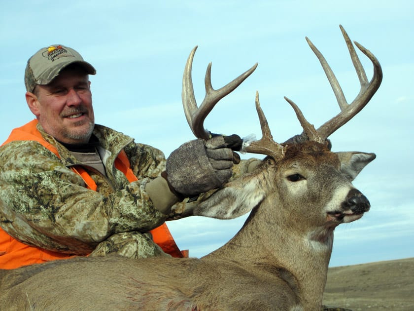 A hunter in camouflage and an orange vest poses with a harvested whitetail deer.