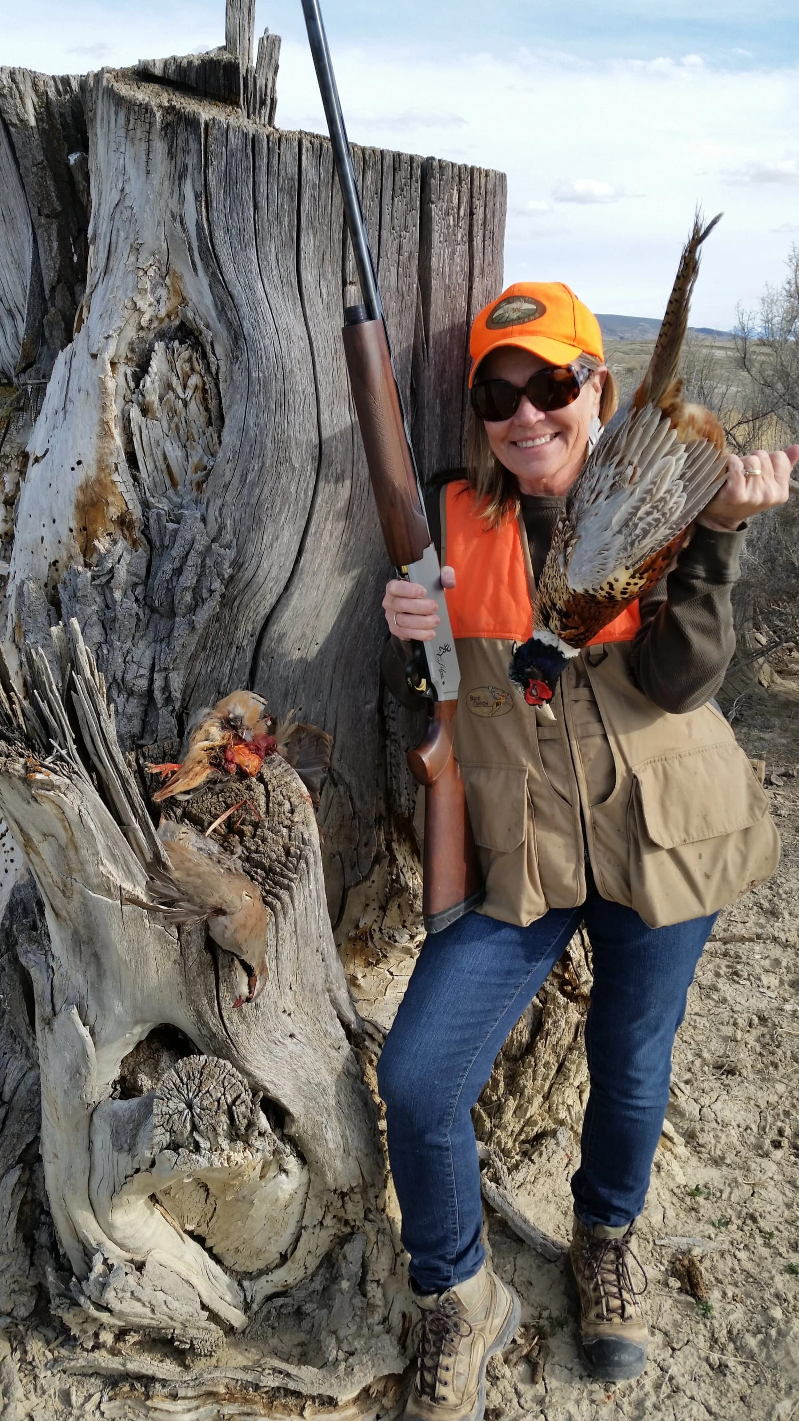 A woman in hunting gear poses with a harvested ring-necked pheasant and chukar partridge next to a tree stump.