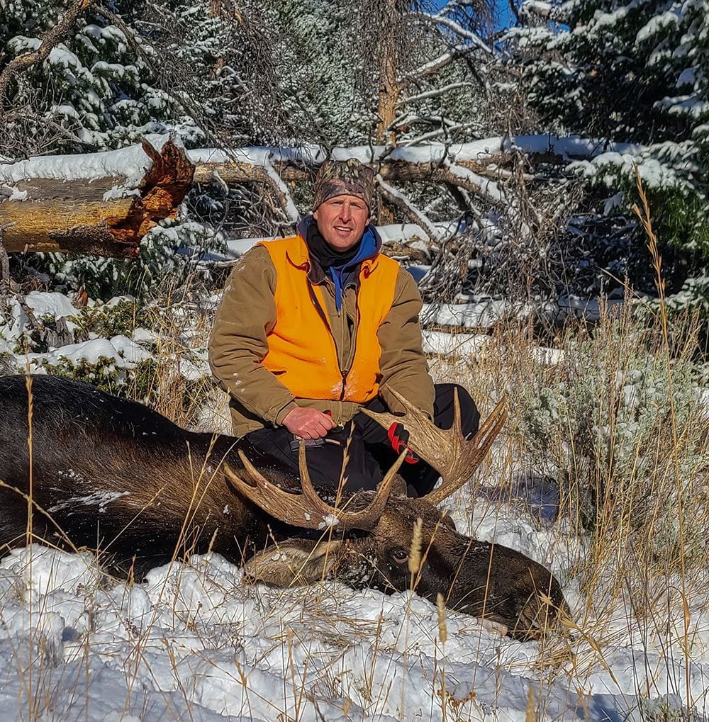 A man in an orange vest poses with a harvested moose in a snowy, wooded area.
