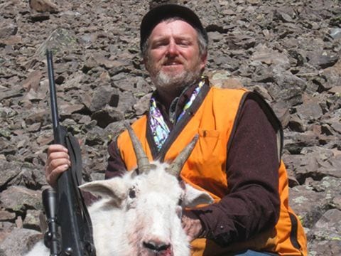 A hunter poses with a harvested mountain goat on a rocky hillside.