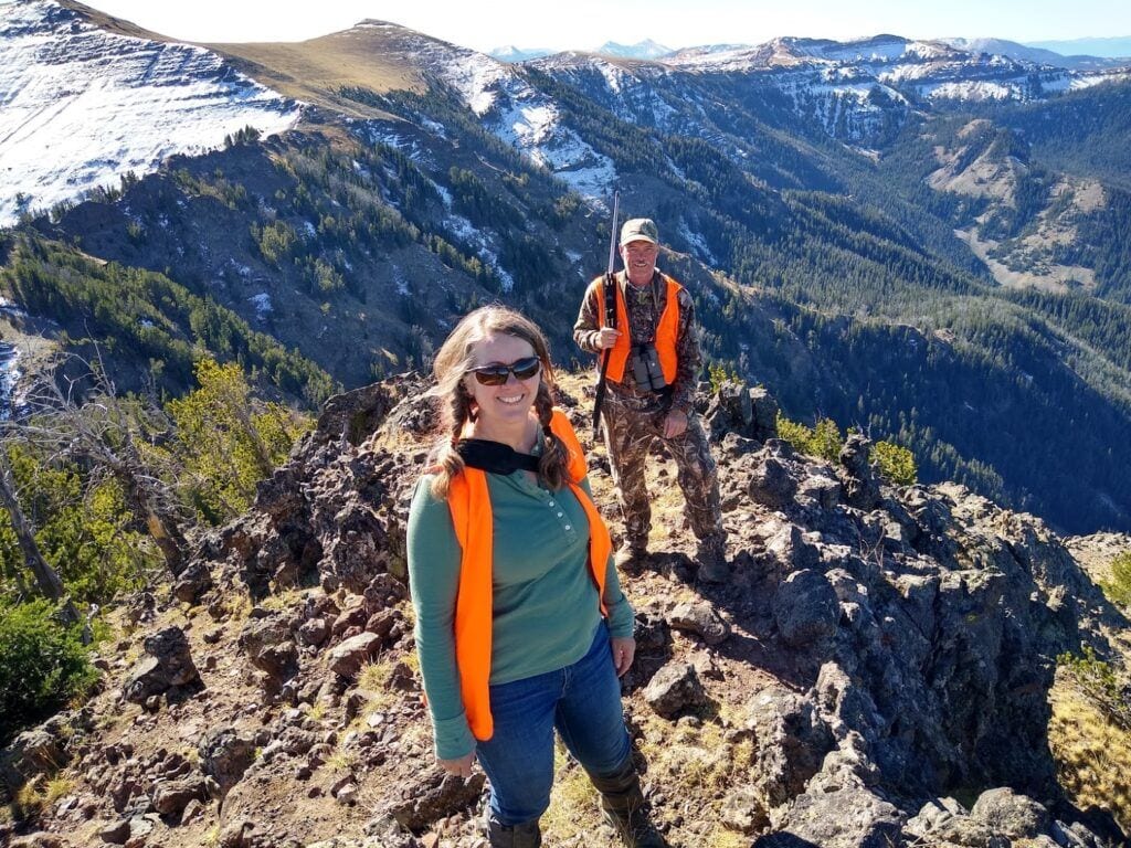 Two hunters in orange vests stand on a rocky mountain ridge with snow-capped peaks and forests in the background.