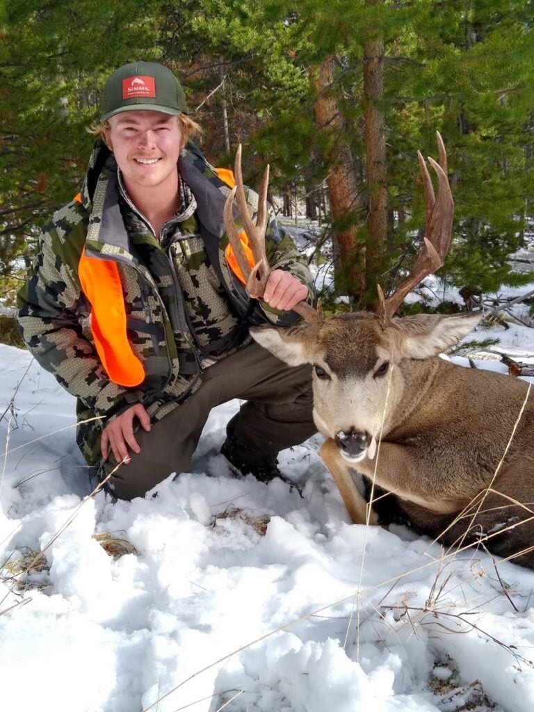 A hunter kneels in the snow next to a harvested deer in a forest.