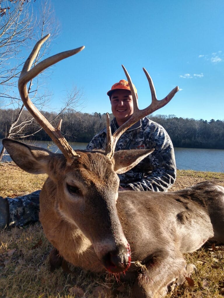 A hunter in camouflage and an orange hat poses with a harvested whitetail deer next to a body of water.
