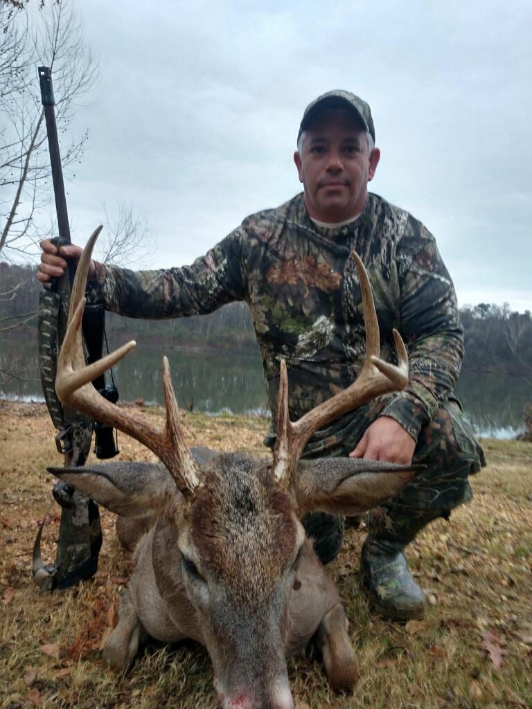 A man in camouflage poses with a harvested whitetail deer and a rifle next to a body of water.